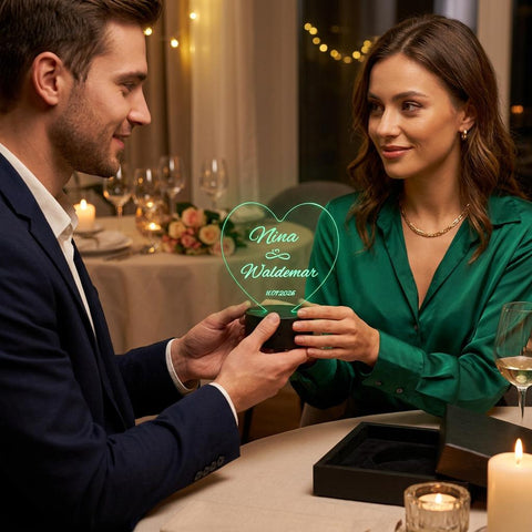 Ein Paar bei einem romantischen Abendessen bei Kerzenschein, bei dem der Mann der Frau eine personalisierte LED-Acryllampe in Herzform mit der Gravur „Nina & Waldemar“ schenkt. Perfektes Hochzeitsgeschenk und Jahrestagsgeschenk.